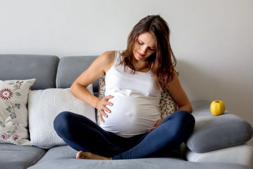Donna in gravidanza seduta a gambe incrociate sul divano che si tiene la pancia